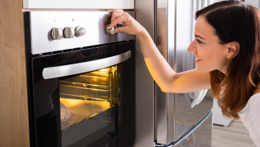 Woman turning a knob on an oven, which has its interior light on and a raw chicken cooking inside.