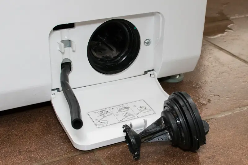 Close-up of a washing machine filter on the floor next to the open panel with the emergency drain hose detached.