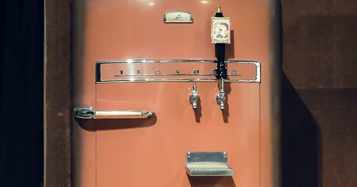 An old peach-colored refrigerator that's been converted into a kegerator, with two taps and a small beer shelf affixed to the refrigerator door.