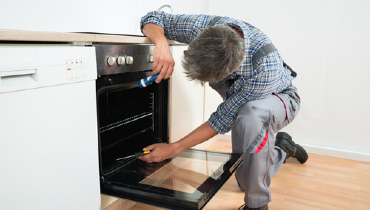 Technician bent over an oven door, shining a flashlight into the back of the oven and holding a screwdriver in his other hand.