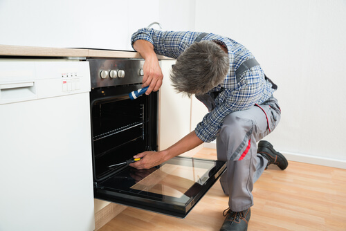 Technician bent over an oven door, shining a flashlight into the back of the oven and holding a screwdriver in his other hand.