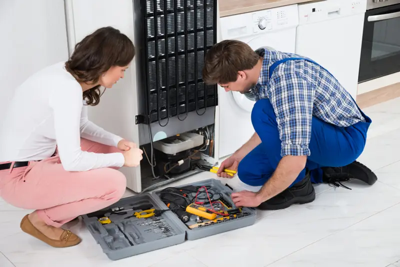 Technician crouched on floor of a kitchen repairing refrigerator; homeowner is crouched down watching him work.