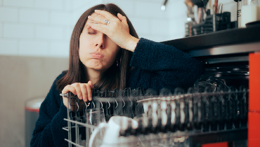 Woman crouched next to open dishwasher, holding on to the top rack, with hand against her forehead making an exasperated face.