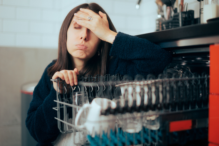 Woman crouched next to open dishwasher, holding on to the top rack, with hand against her forehead making an exasperated face.
