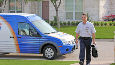 Service technician carrying a tool bag and walking up a sidewalk with work van parked on the street behind him.