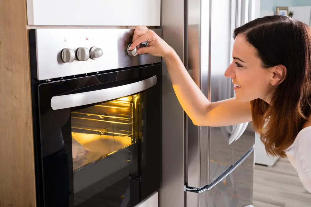 Woman turning a knob on an oven, which has its interior light on and a raw chicken cooking inside.