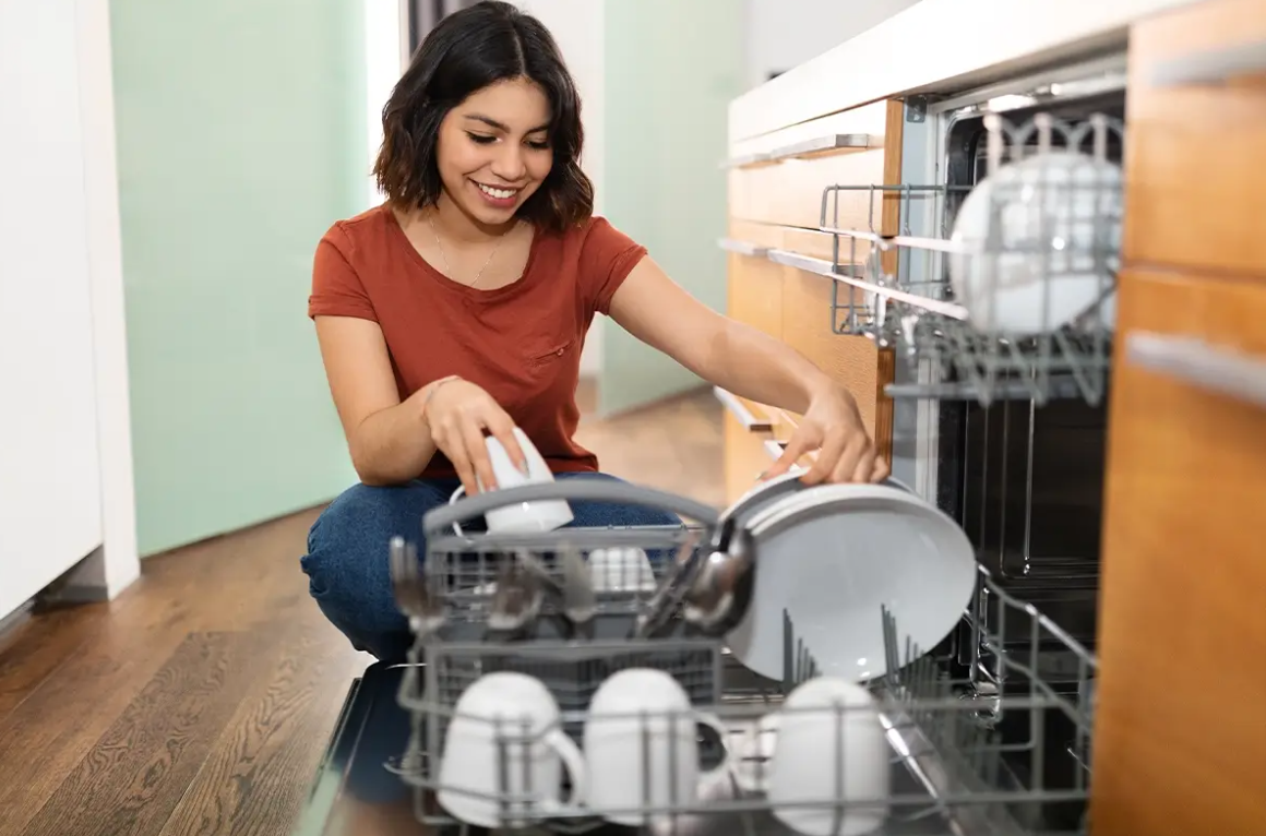 A woman wearing a red shirt and jeans is smiling while placing a mug and plate into a dishwasher. | how-to-load-dishwasher