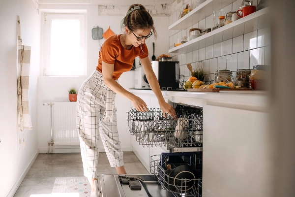 Woman unloads a dishwasher in a small, white kitchen.
