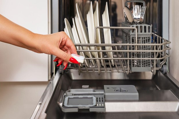 A woman's manicured hand places a tablet into an open dishwasher with dirty dishes on the lower rack.