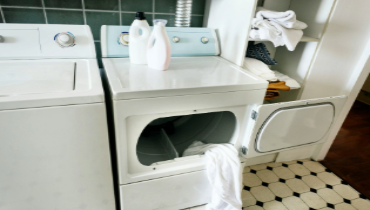 An open, broken-looking white washer in a home, with laundry hanging out the front, that requires a Plano appliance repair solution to fix.