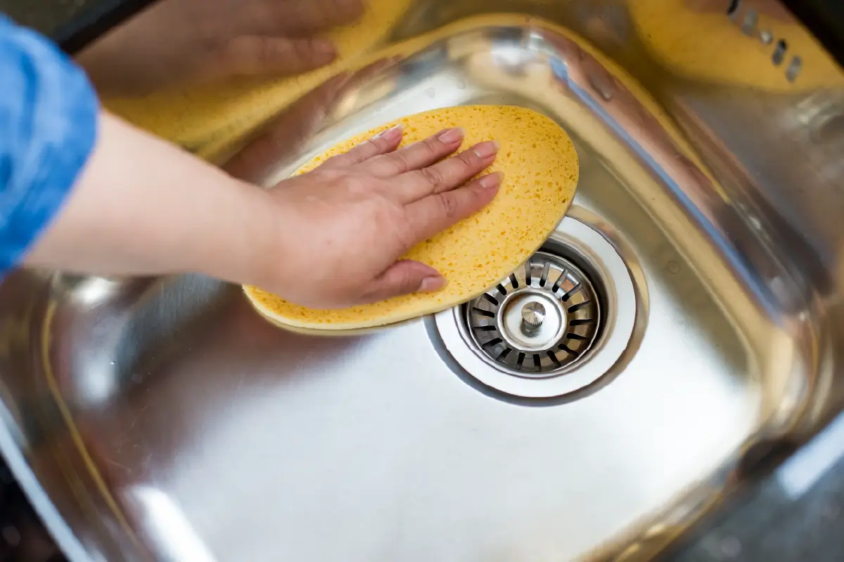 A hand wiping down a stainless steel sink with a yellow sponge.