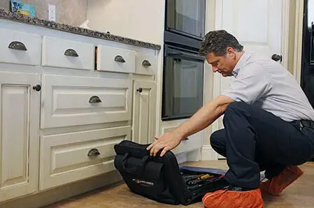 Mr. Appliance technician in orange booties unpacking tool bag on floor of residential kitchen.